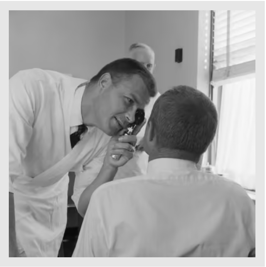 black and white image of a mid 20th century White male doctor using an otoscope to look into a male patient's eye
